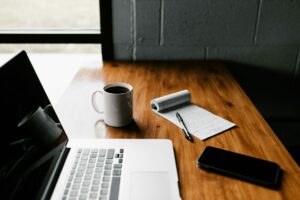 A remote workspace setup with a laptop, a cup of coffee, a notepad, a pen, and a smartphone placed on a wooden desk.