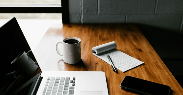 A remote workspace setup with a laptop, a cup of coffee, a notepad, a pen, and a smartphone placed on a wooden desk.